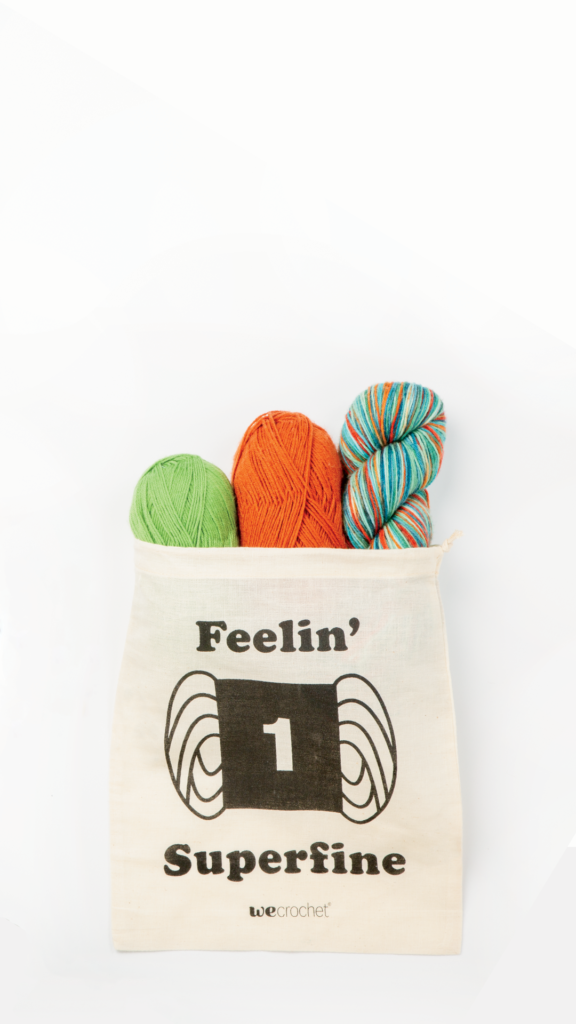 A cream-colored project bag that says "Feelin' Superfine" with a yarn weight 1 icon, filled with colorful yarn.