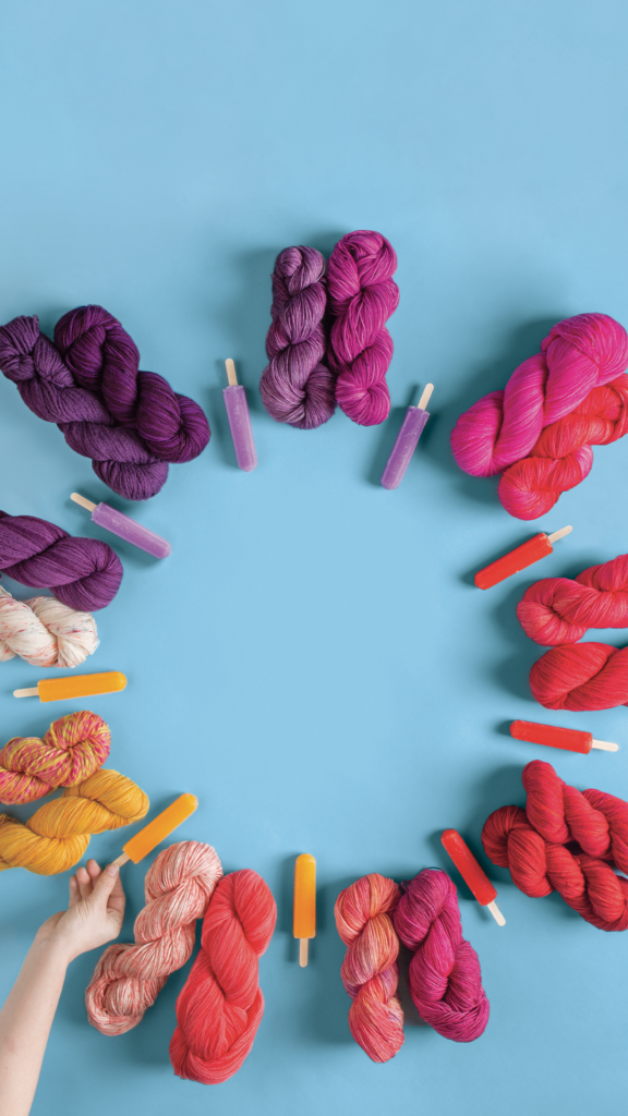 On a blue background, a ring of colorful yarn ranging from purple to pink to red to orange along with matching popsicles.