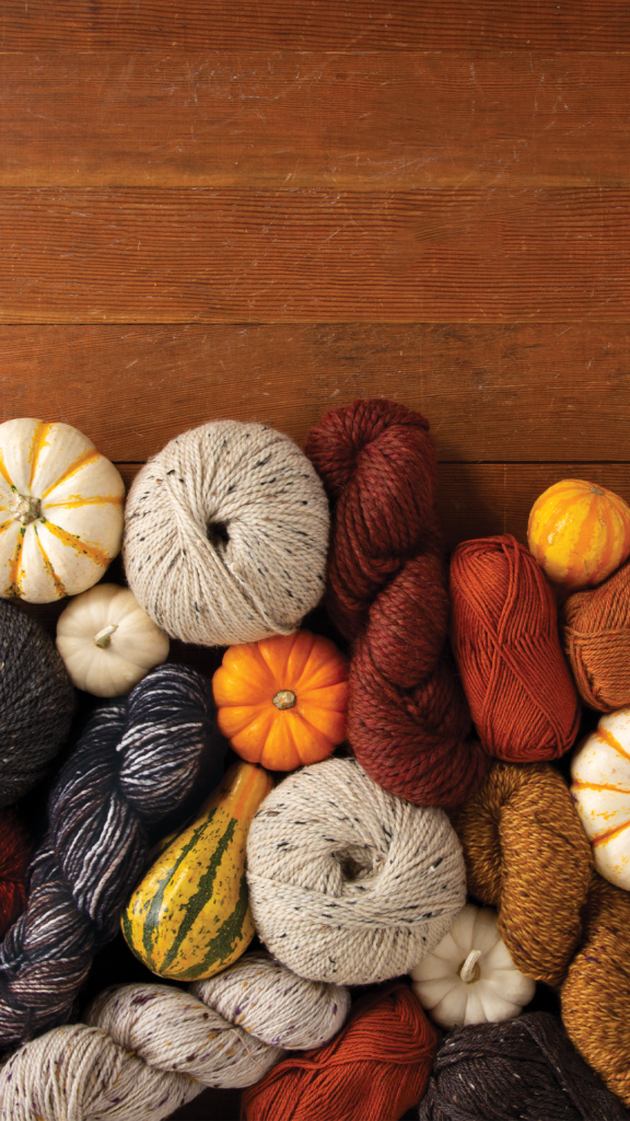 A large pile of various yarn in fall colors (rust, pumpkin, gray, maroon), along with assorted decorative pumpkins.