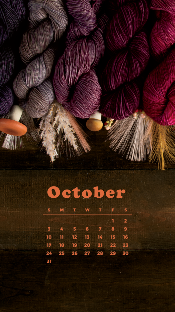 An October 2021 calendar on a dark wood background with assorted pink and purple hanks of yarn decorated with mushrooms and wheat.
