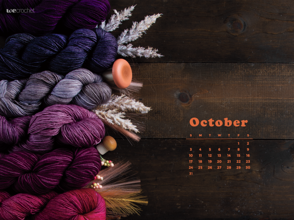 An October 2021 calendar on a dark wood background with assorted pink and purple hanks of yarn decorated with mushrooms and wheat.