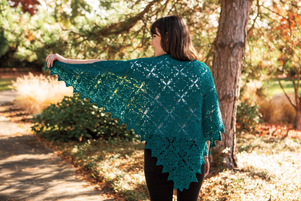 The Placid Shawl is a teal colored shawl made of square motifs, joined into a triangular shape. A model shows the shawl with its wingspan spread out.