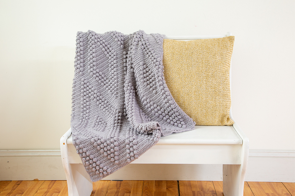 A gray textural crocheted blanket draped on a white bench