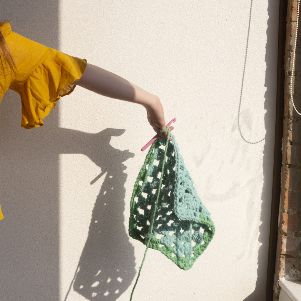 a hand holds an in-progress crochet granny square and casts a shadow on the wall