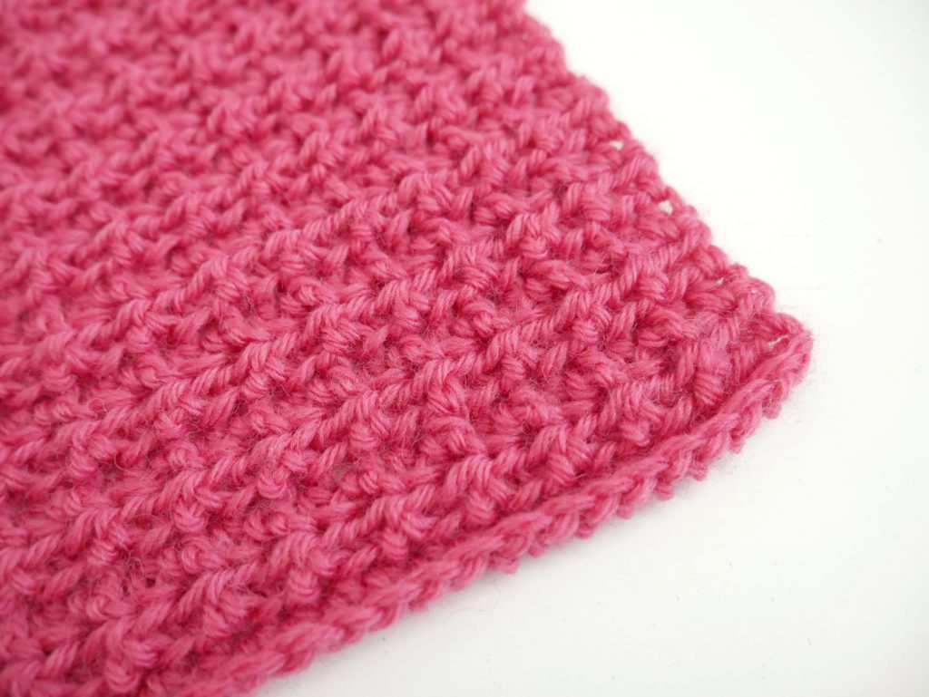 The corner of a bright pink crochet swatch with a waffle-ish texture on a white background.