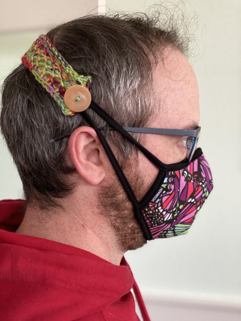 A man's head in profile, demonstrating the use of Ear Savers (a strap with a button), wrapped around his head and connecting to the straps of a protective face mask made of colorful fabric.