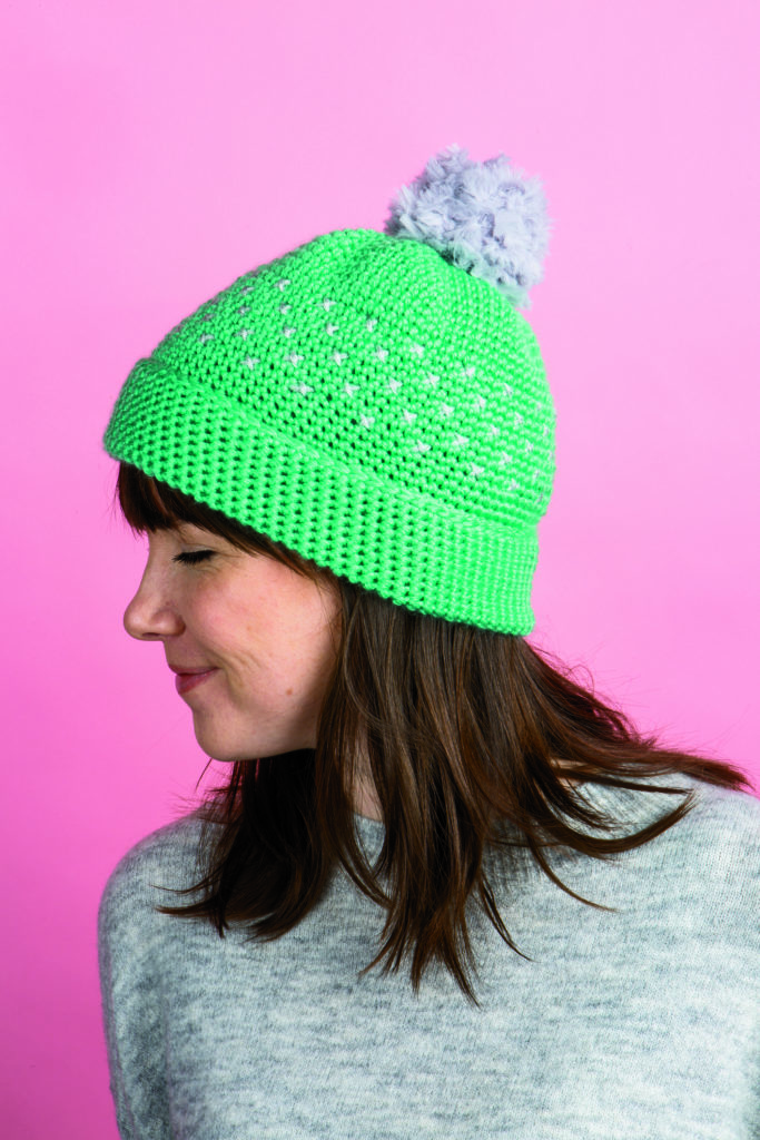 A model wears a green crocheted hat with a gray pom-pom
