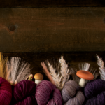 A dark wood background with assorted pink and purple hanks of yarn decorated with mushrooms and wheat.