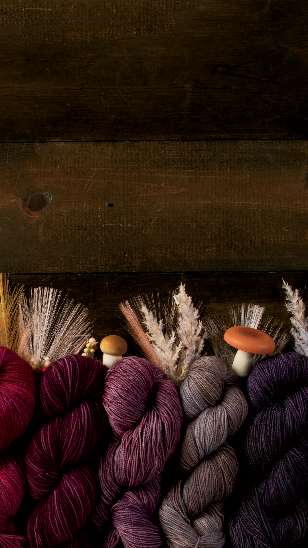 A dark wood background with assorted pink and purple hanks of yarn decorated with mushrooms and wheat.